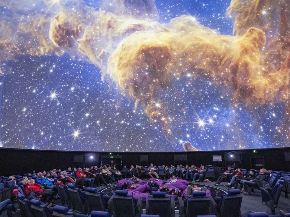 Zuschauer in einem Planetarium blicken auf eine kosmische Darstellung an der Kuppeldecke.