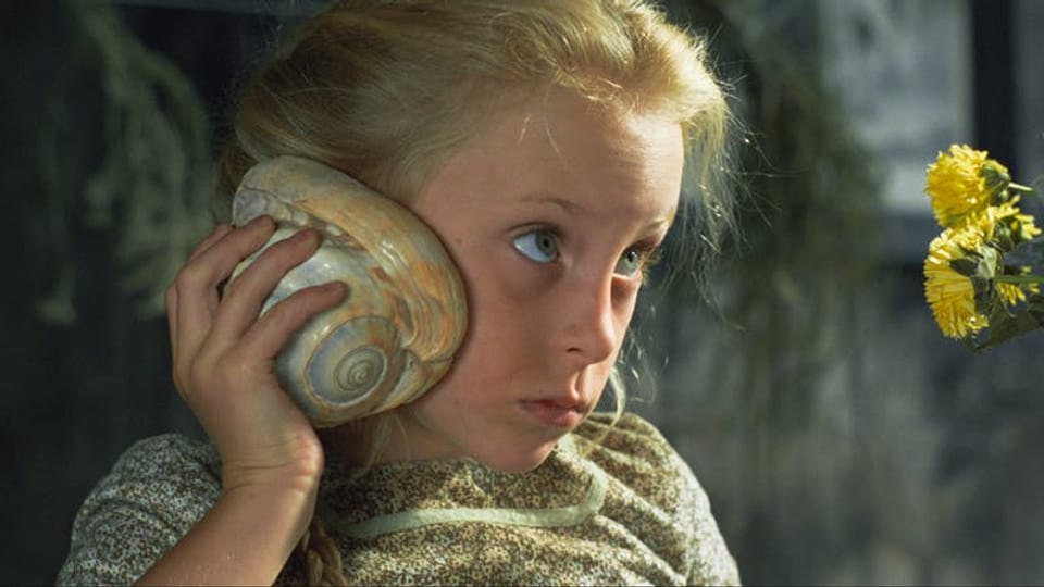 Ein blondes Mädchen hält eine Muschel an sein Ohr.