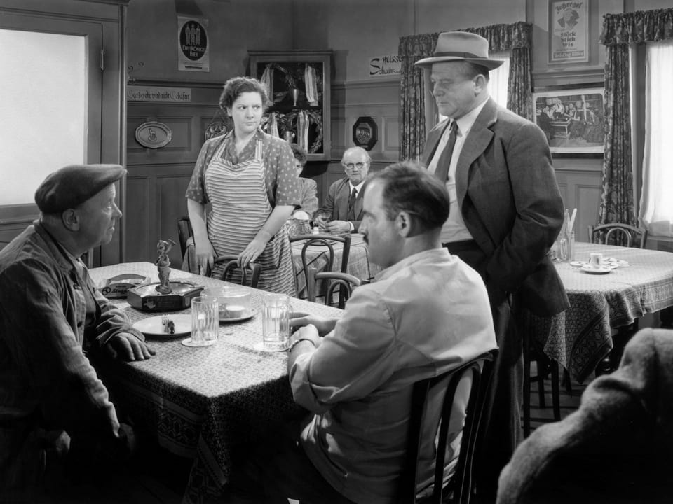 Zwei Männer am Tisch in einem Gasthaus, davor stehen ein Mann und eine Frau.