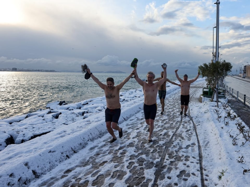 Vier Männer in Badehosen rennen am Meer eine schneebedeckte Strasse entlang.