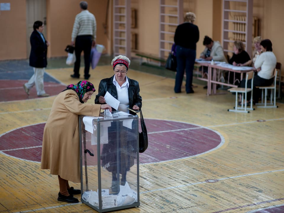 Zwei Frauen legen ihren Wahlzettel in die Urne. Das ganze findet in einer Turnhalle statt.
