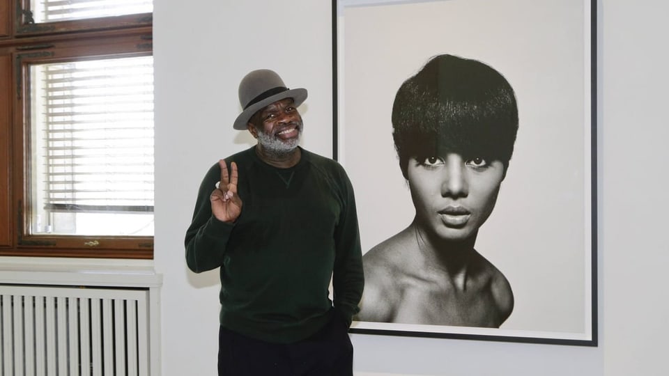 Ein schwarzer Mann posiert vor einer schwarz-weiss Fotografie.
