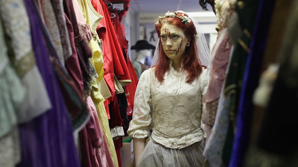 Eine Dame im Brautkleid mit künstlichem Blut im Gesicht steht zwischen Kostümen