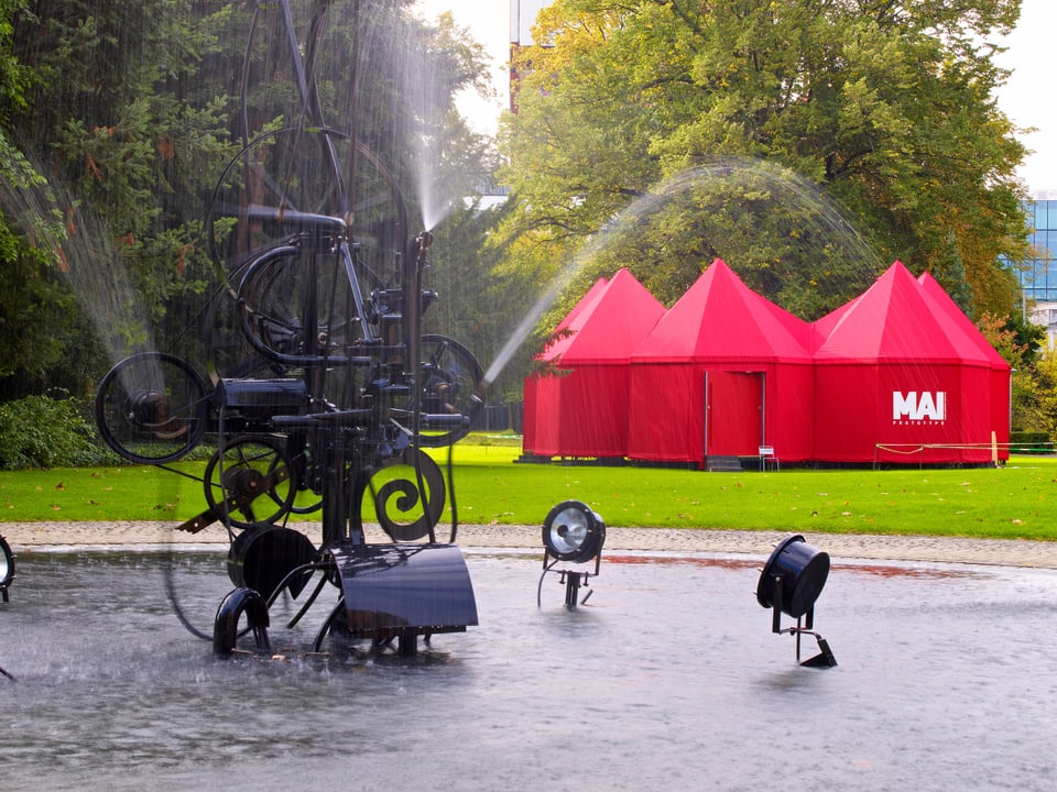 Rote aneinandergelegte Zeltpavillons auf einer Wiese, davor der Brunnen vor dem Museum Tinguely.