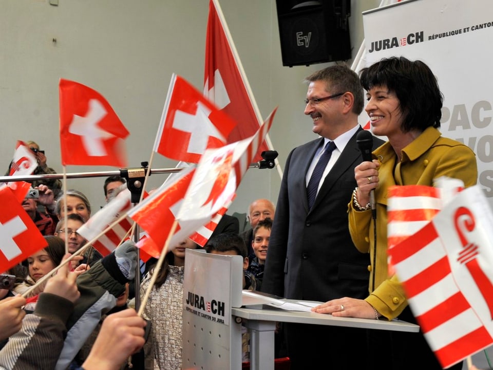 Mann und Frau am Rednerpult. Darumherum Jura- und Schweizerflaggen.
