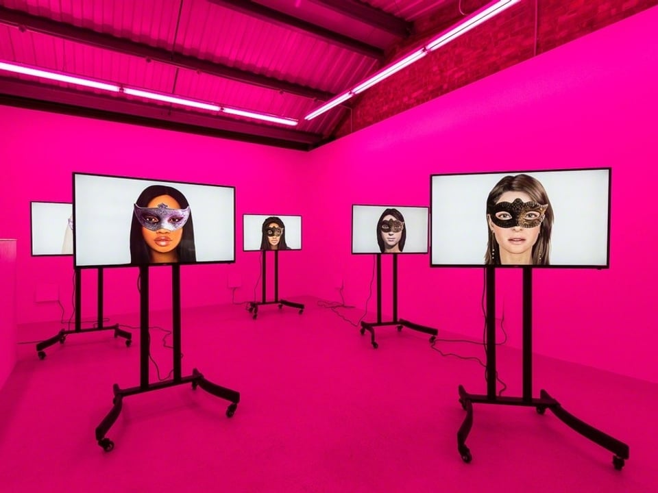 Ein rosa erleuchteter Raum mit fünf Bildschirmen, darauf sind Avatare von Frauen mit Augenmasken zu sehen.