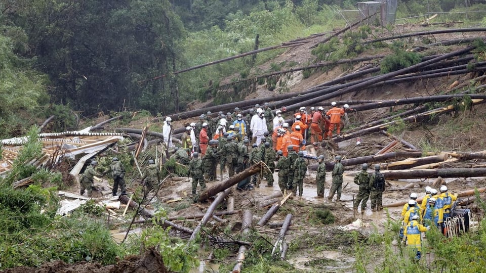 Umgestürzte Bäume an einem Abhang, darin stehen weiss und orange gekleidete Helfer zusammen mit Soldaten.