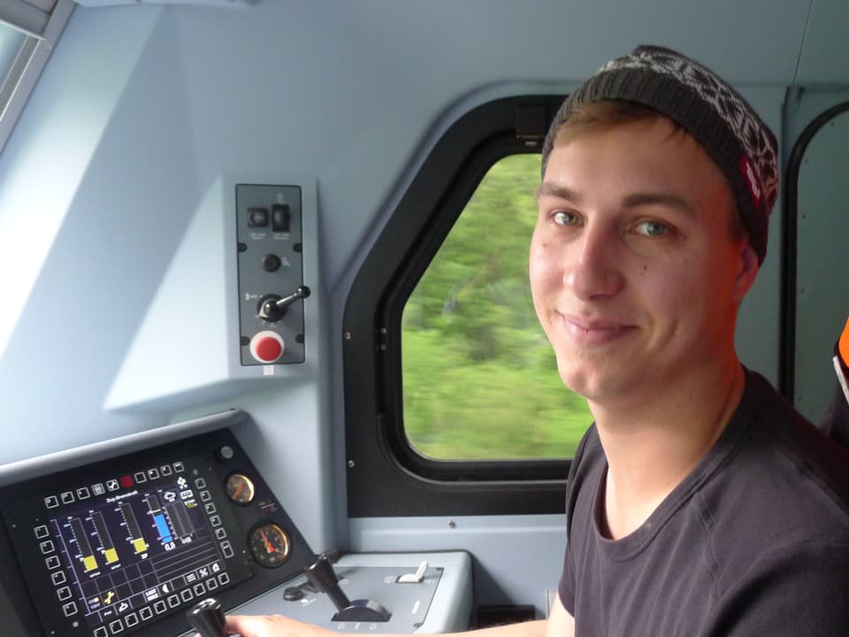 Der junge blondharige Lokführer mit einer Kappe auf dem Kopf sitzt vorne im Führerstand der Lokomotive.