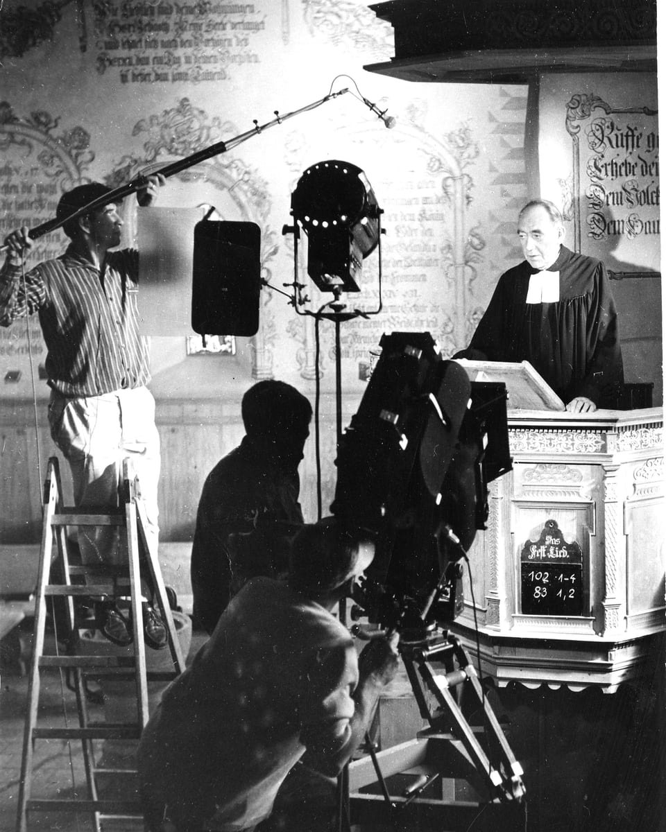 Ein Pfarrer steht in der Kanzel einer Kirche und predigt. Unterhalb vor ihm steht die Kamera. Ein Filmscheinwerfer wirft Licht auf ihn. Links im Bild auf einer Bockleiter steht der Tonangler mit der Tonangel.