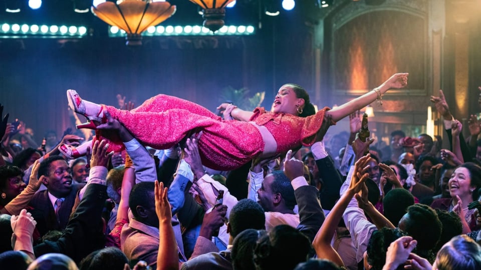 Filmszene in einem Nachtclub: Eine Frau mit Kleid und hohen Schuhen wird von der Menge auf Händen getragen.