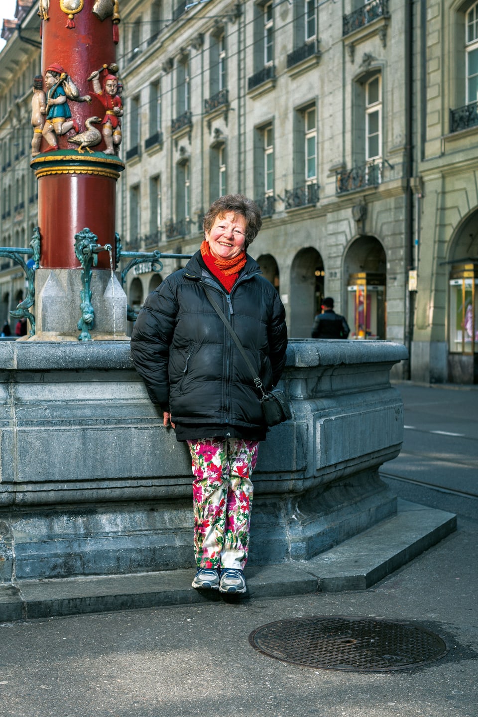 Eine Frau lehnt an einen Brunnen in der Berner Altstadt, sie trägt eine schwarze Dauenejacke und geblümte Hosen. 
