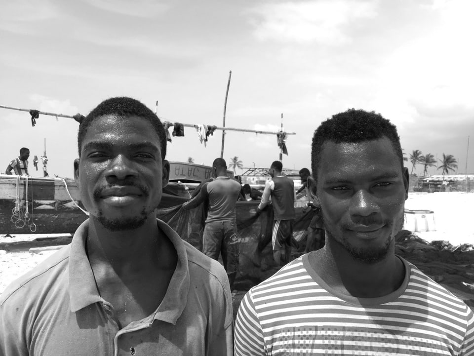 Schwarz-Weiss-Aufnahme: Porträtaufnahme der beiden Männer. Im Hintergrund hängen Fischer ein langes Netz an ein Boot.