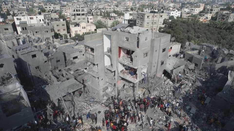 Ein Bild von einem zerstörten Wohnblock. Menschen stehen vor dem Gebäude.