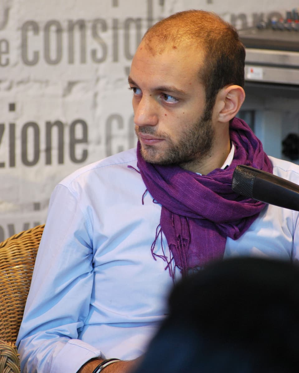 Giovanni Tiziano mit dem Blick von der Kamera abgewandt sitzt vor einem Mikrophon in einem Sessel.