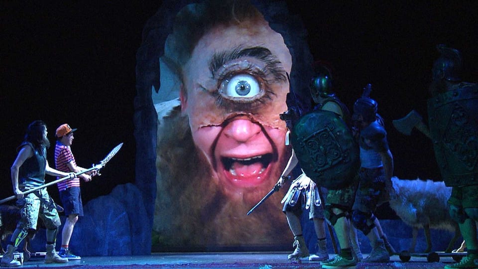Eine Szene aus der Theateraufführung, ein riesiger Zyklopenkopf und Männer die sich vor ihm fürchten.