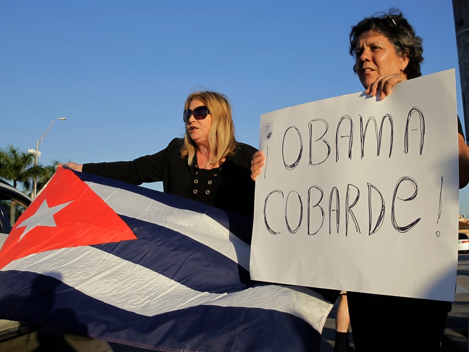 Eine Frau (links) hält die kubanische Flagge, eine weitere Frau (rechts) hält ein Schild mit der Aufschrift «Obama Cobarde (Feigling)»»