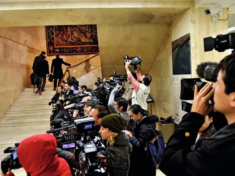 Ein paar Konferenzteilnehmer gehen eine Treppe hoch, rechts im Bild stehen unzählige Journalisten mit Kameras.