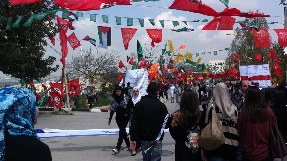 Mehrere Studenten, junge Frauen und Männer, laufen über den Campus. Über ihnen hängen bunte Fahnen und die Flagge Palästinas.