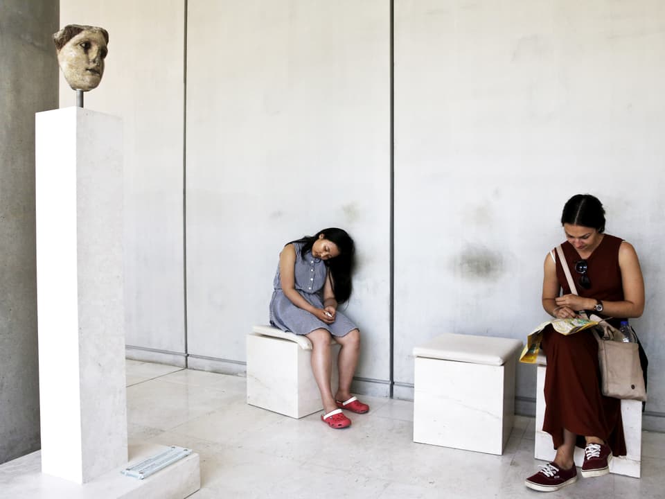 Zwei Besucherinnen ruhen sich auf Hockern aus, neben einer antiken Statue.