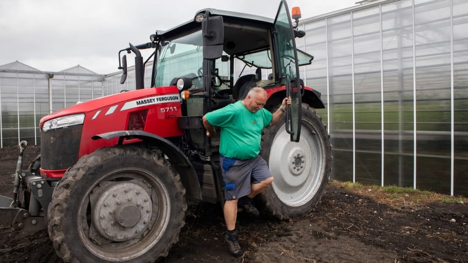 Mann in grünem T-Shirt steigt aus rotem Traktor, dahinten weisses Gewächshaus.