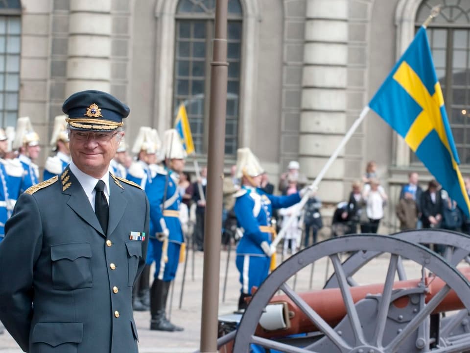 König Carl Gustaf in Uniform mit Hut lächelnd vor einer Parade stehend.