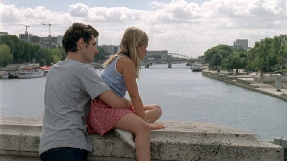 Junger Mann steht auf einer Brücke, ein Mädchen sitzt vor ihm auf dem Geländer, das eine Steinmauer gleicht.
