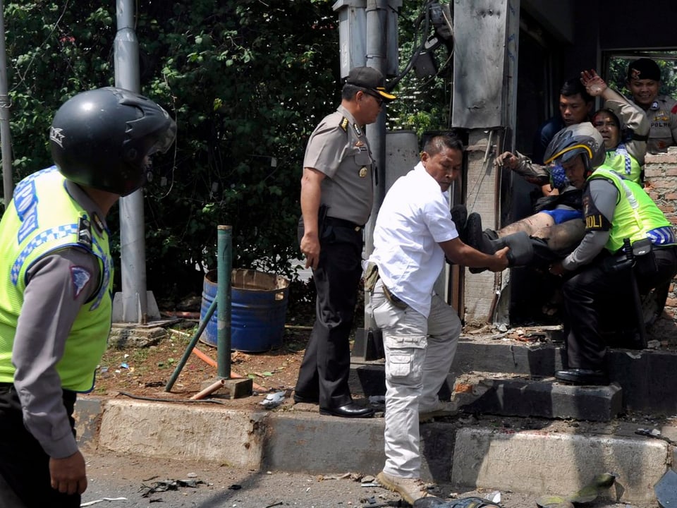 Polizisten und ein weiss gekleideter Sanitäter tragen einen Verletzen aus einem Haus.