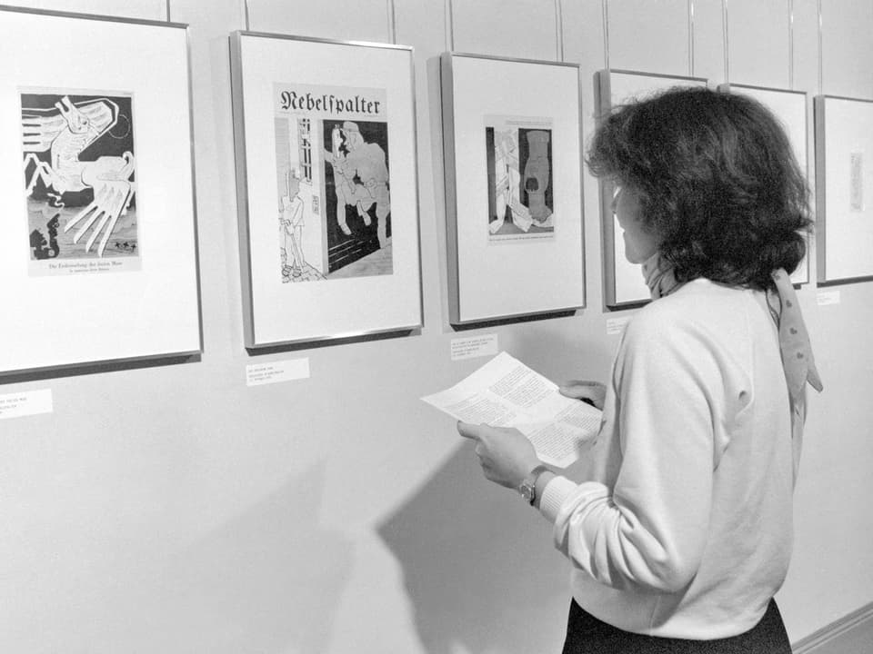 Schwarzweissphoto: Eine Frau betrachtet Bilder von satirischen Arbeiten.
