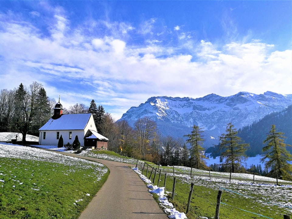 Ein Weg führt zur Kapelle, auf den Wiesen daneben liegt noch ein wenig Schnee. Im Hintergrund sind schneebedeckte Berge vorhanden.