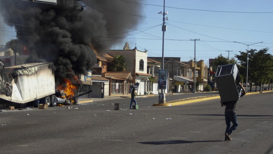 Links ist brennender Lastwagen auf einer Strasse zu sehen. Rechts davon tragen Leute Objekte herum.
