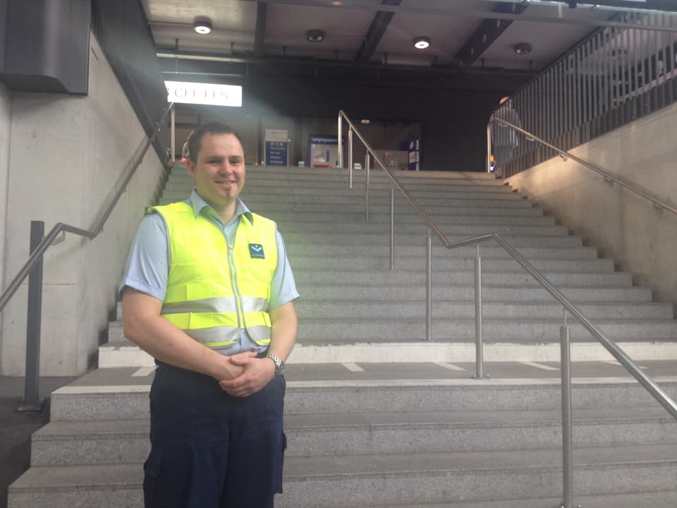 Martin Jeker steht mit gelber Weste vor der Treppe, die Hände im Schoss gefaltet, mit Spitzbart. 