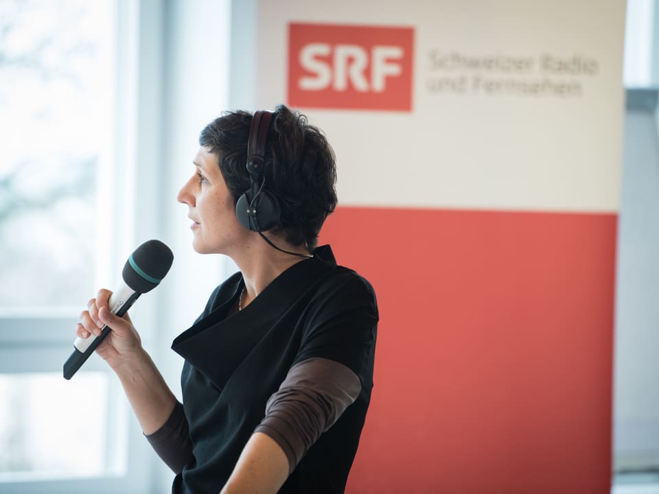 Frau mit Mikrophon blickt zum Fenster hinaus, dahinter das SRF-Logo.