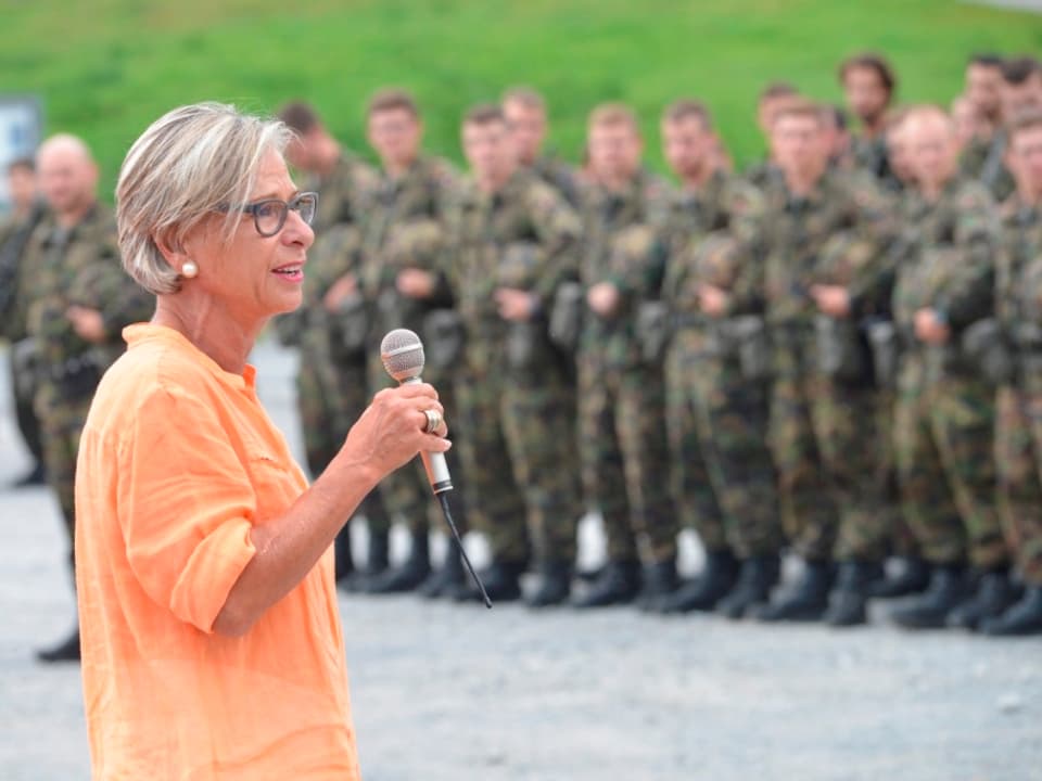 Eine Frau mit oranger Bluse und Mikrofon in der Hand steht vor Männern in Uniform.
