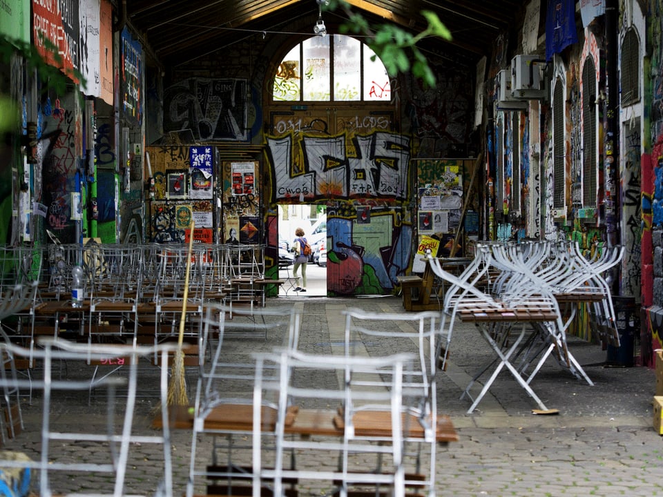 Blick in die Reitschule Bern: Klappstühle, Wände mit Graffiti, im Hintergrund geht eine Frau durch eine Tür.