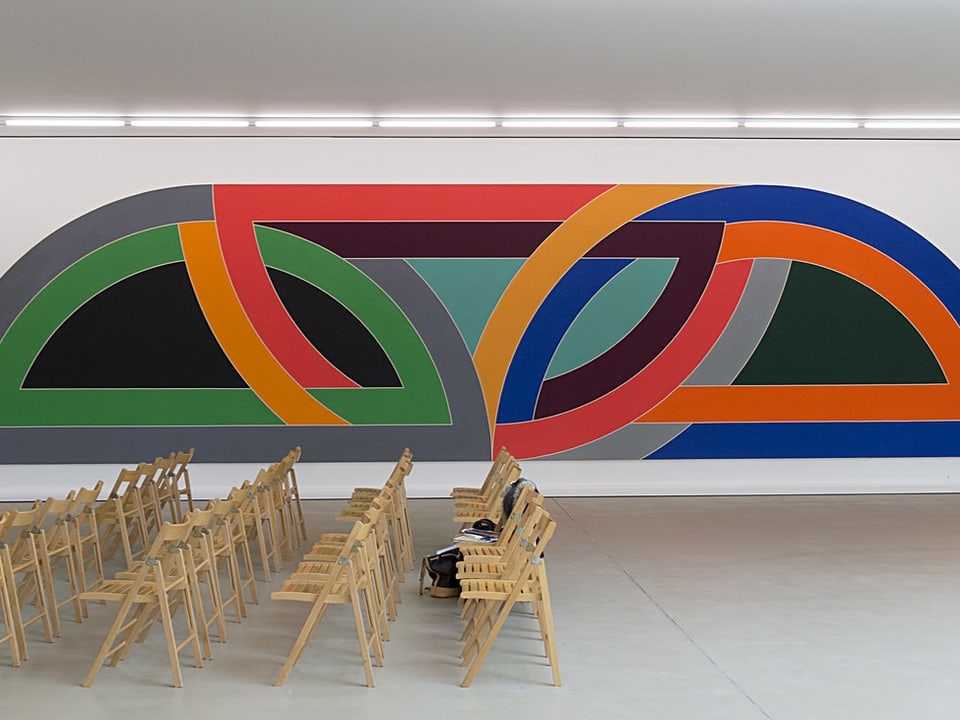 Ein farbiges, geometrisches Bild (mit farbigen Bögen) auf weisser Wand, davor Holzstühle.
