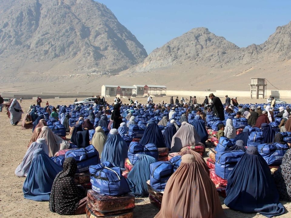 Afghan families sit with their backs to the camera on sandy ground in the desert.