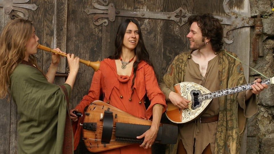 Drei Musiker in mittelalterlichen Kostümen vor einem alten Tor.