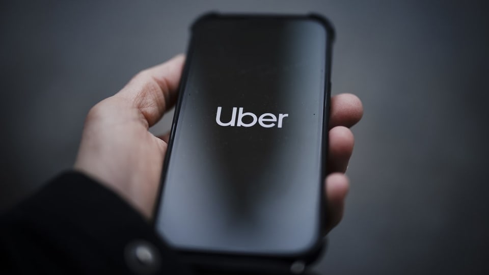 Ein Smartphone in einer Hand zeigt das Uber-Logo in weisser Schrift auf schwarzem Hintergrund