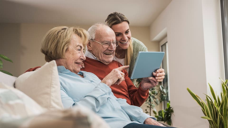 Älteres Paar und junge Frau betrachten gemeinsam ein Tablet auf dem Sofa.