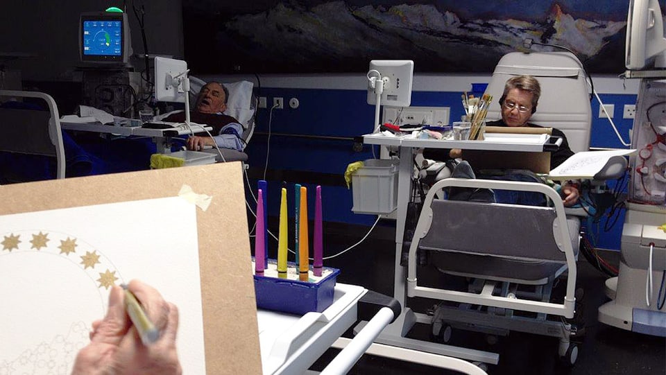 Blick in die Dialysestation über die Schulter einer malenden Person hinweg zu einer anderen, die ebenfalls kreativ tätig ist.