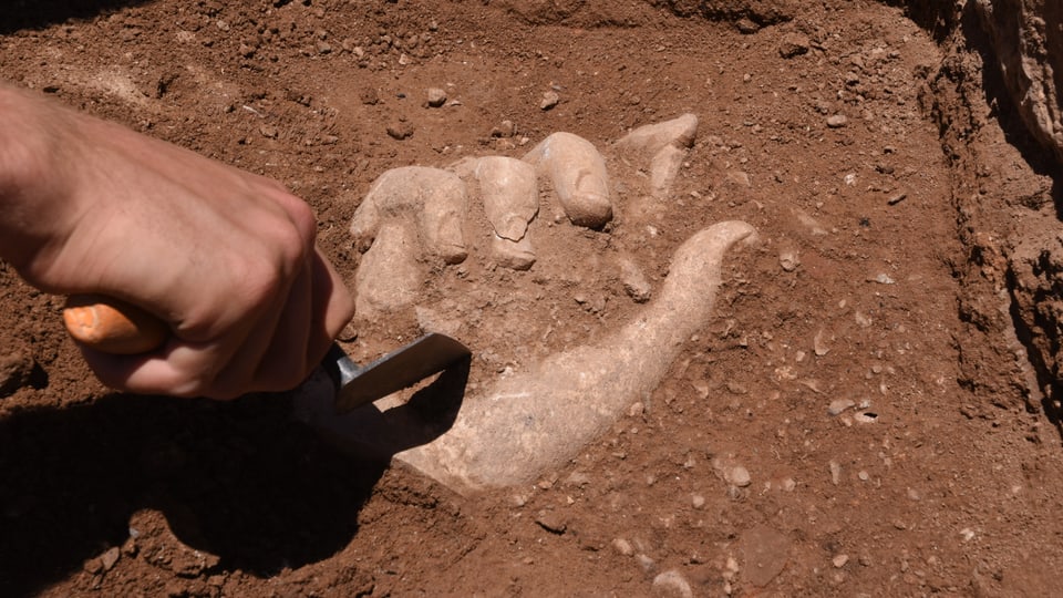 Eine Hand mit Schaufel gräbt eine Hand aus Stein aus.