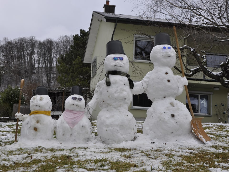 Eine Schneemann-Familie mit vier Schneemännern steht im praktisch schneefreien Garten vor einem Haus.