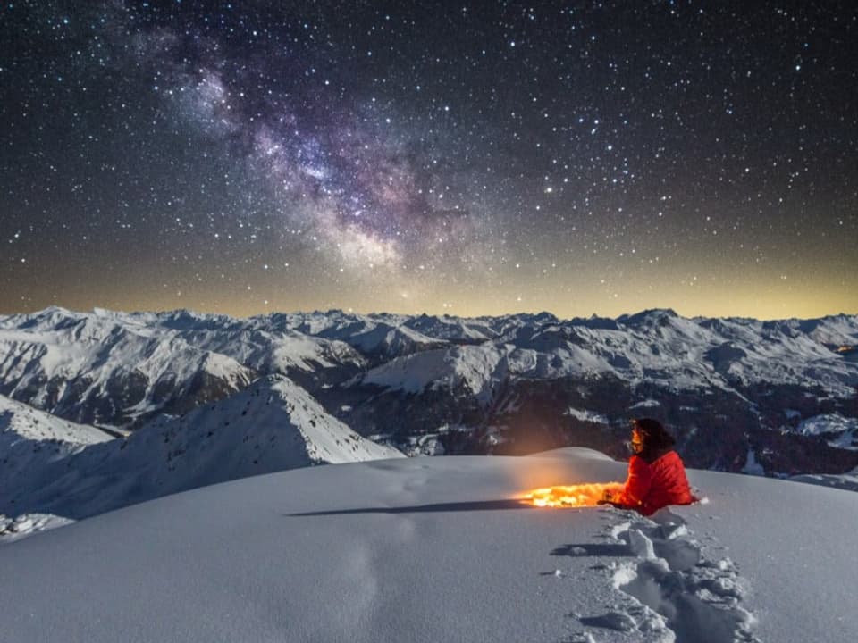 Mensch an Feuer im Schnee, Blick auf verschneites Alpenpanorama und viele Sterne am Himmel.