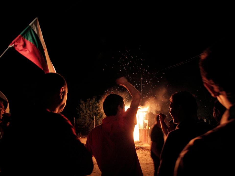 Menschen mit Flagge stehen in der Dunkelheit vor einer brennenden Hütte.