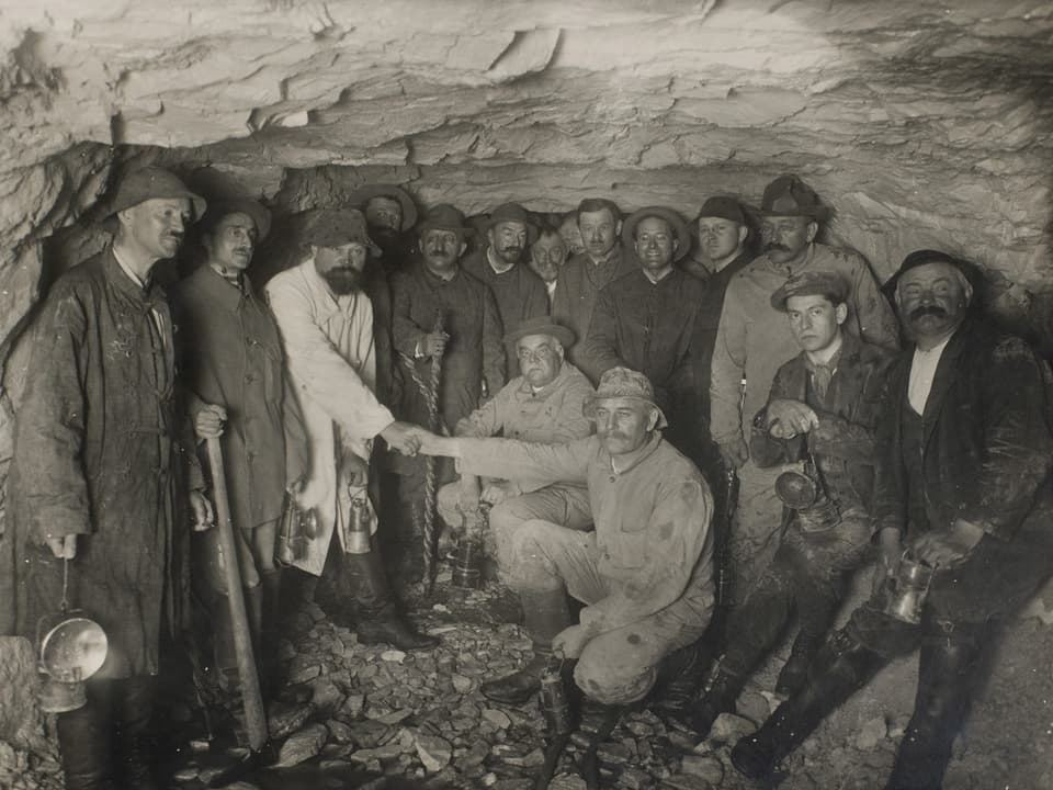 Schwarz-Weiss-Fotografie von Arbeitern in einem Tunnel, zwei geben sich die Hände