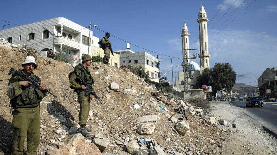 Bewaffnete Soldaten vor einem Haus, im Hintergrund die Minarette einer Moschee.