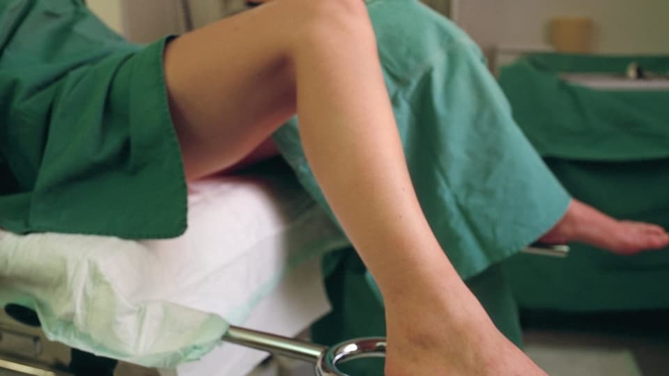 Eine Frau sitzt auf einem Stuhl bei einem Frauenarzt, sie trägt ein grünes Tuch