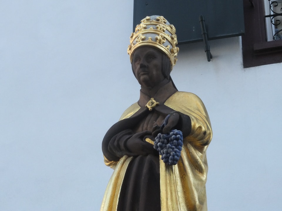 Figur aus rotem Stein, trägt Hut mit goldenen Verzierungen und einen goldenen Mantel, dazu hält er Trauben in der Hand. 