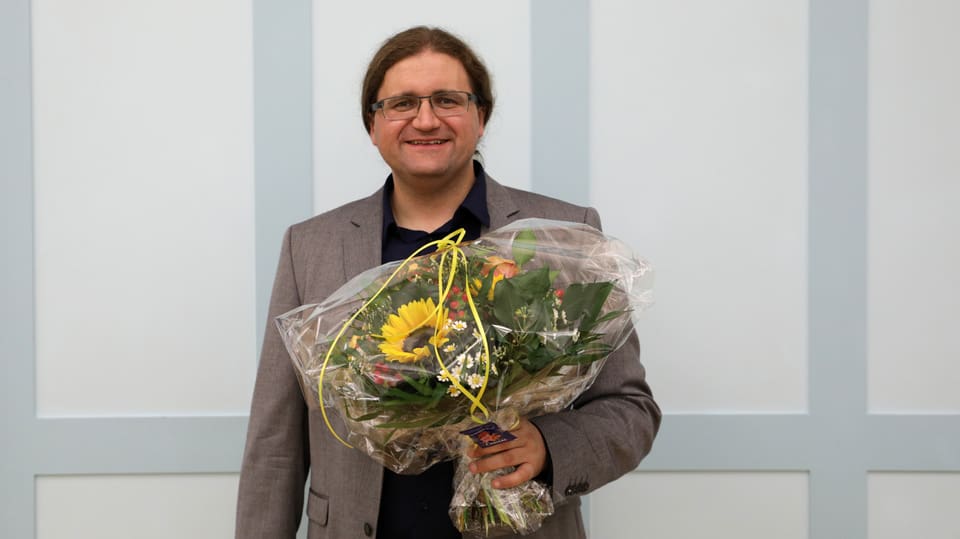 Ein Mann mit Brille steht vor einer Wand und hält einen Blumenstrauss in der Hand. 