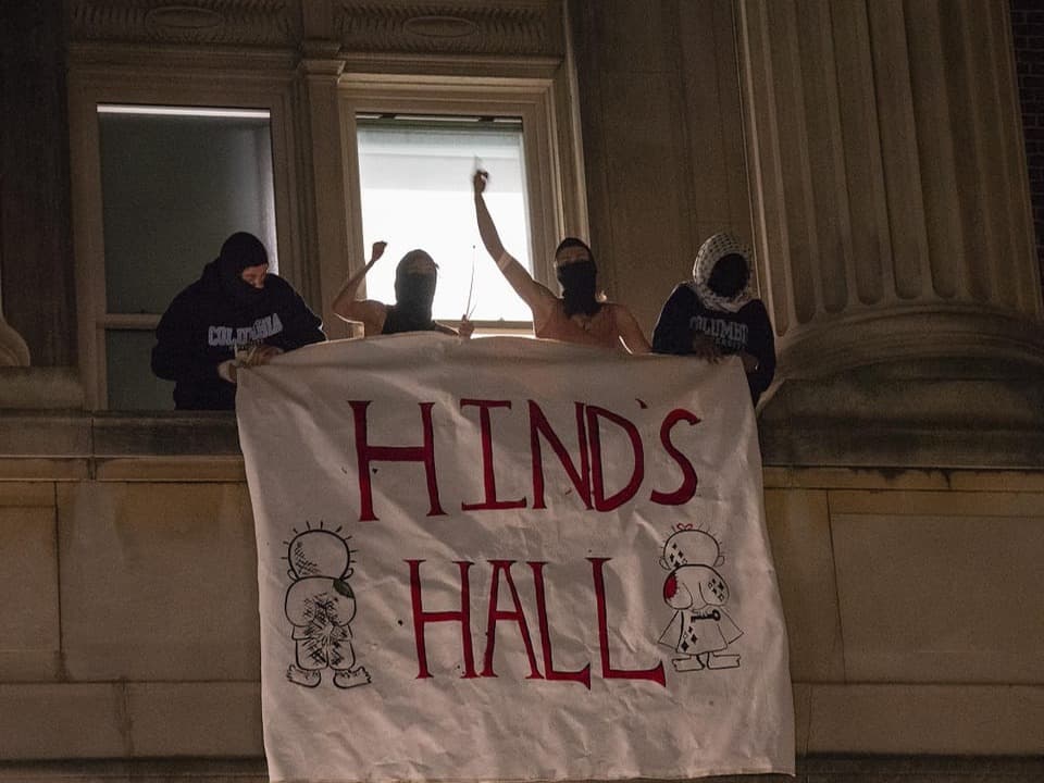 Personen mit Masken halten ein Banner mit der Aufschrift 'HINDS HALL' an einem Gebäude.
